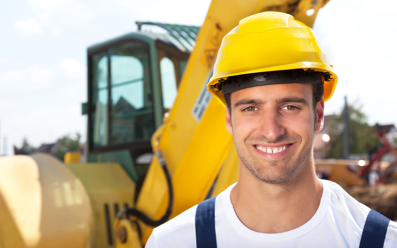 Bauarbeiter mit gelbem Schutzhelm lächelt vor einer gelben Baumaschine auf der Baustelle.