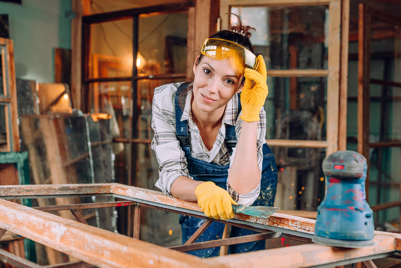 Betriebshaftpflicht Handwerker Handwerkerin mit Schutzbrille und Handschuhen restauriert einen alten Holzrahmen in der Werkstatt.