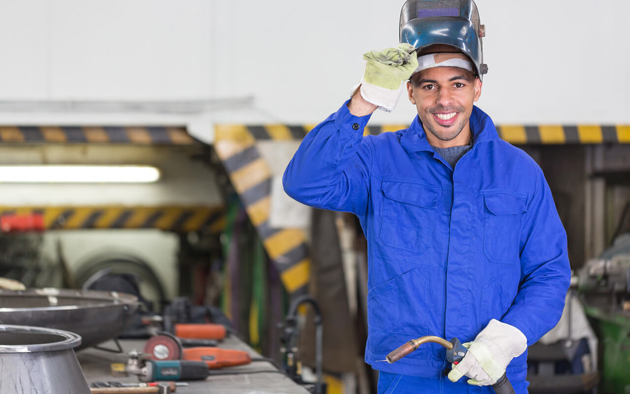Schweißer in blauer Schutzkleidung hebt seinen Helm und lächelt, während er den Schweißbrenner in der Werkstatt hält.