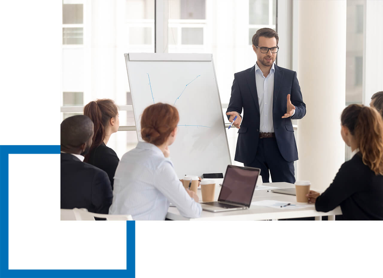 Geschäftstrainer im Anzug präsentiert an einem Flipchart vor Mitarbeitenden, die mit Laptops und Kaffeebechern am Konferenztisch sitzen.