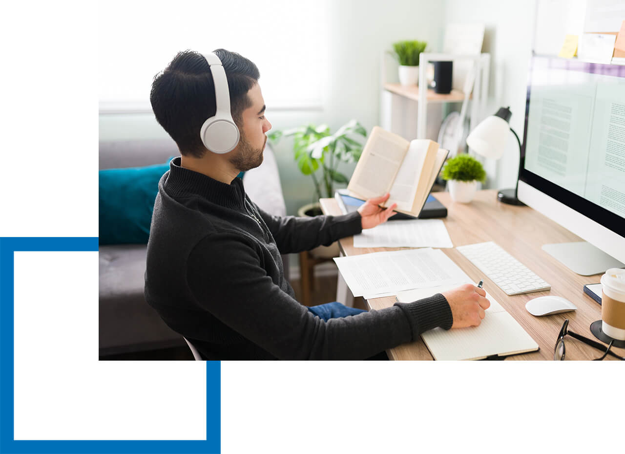 Mann mit Kopfhörern liest Buch und macht Notizen am Schreibtisch mit Monitor.