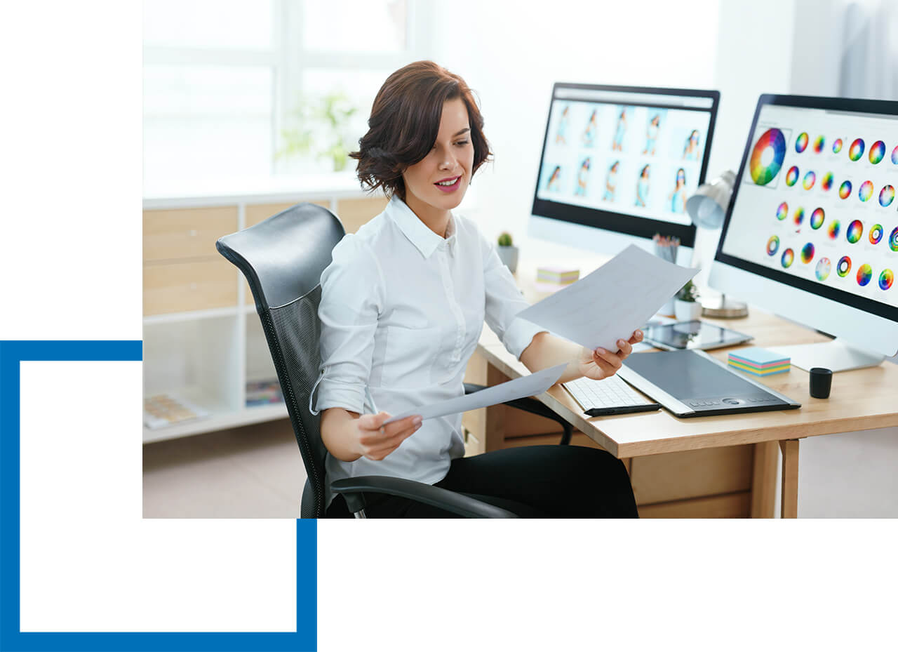 Warum ist sie für Webdesigner wichtig? Grafikdesignerin überprüft Ausdrucke, während auf zwei Monitoren Layouts und Farbpallets angezeigt werden.