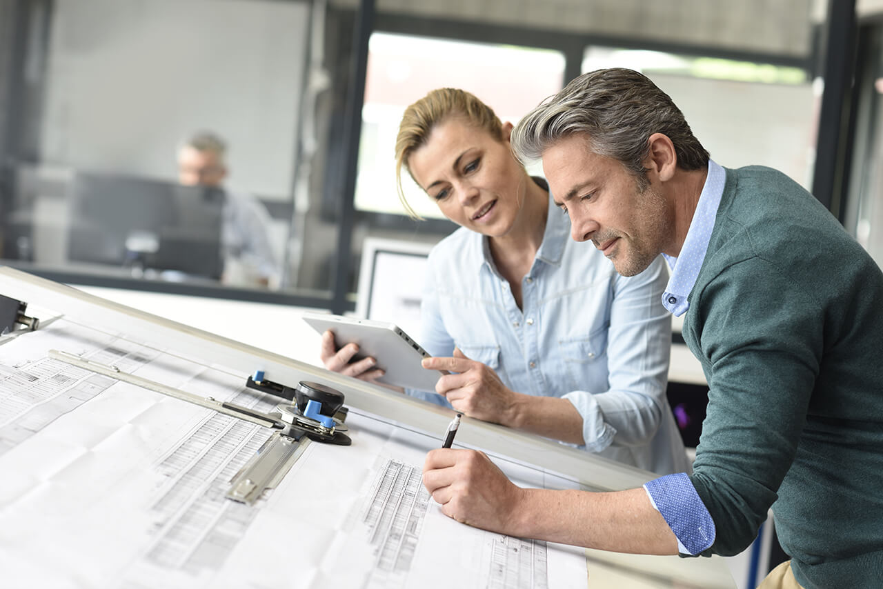 Berufshaftpflicht für Architekten Zwei Personen analysieren gemeinsam technische Zeichnungen mit Tablet und Zeichenwerkzeug.