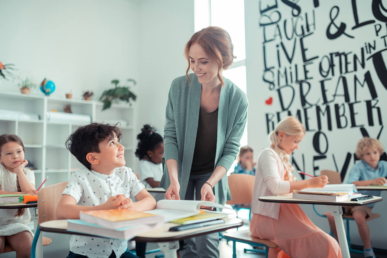 Lehrerin hilft einem lächelnden Schüler an seinem Tisch, während andere Grundschulkinder im kreativ gestalteten Klassenraum arbeiten.