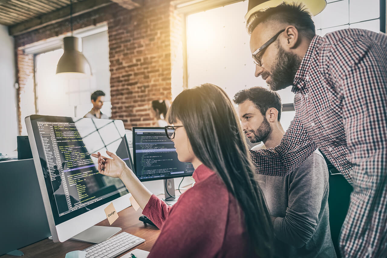 Team bespricht Programmcode gemeinsam vor mehreren Monitoren im modernen Büro.