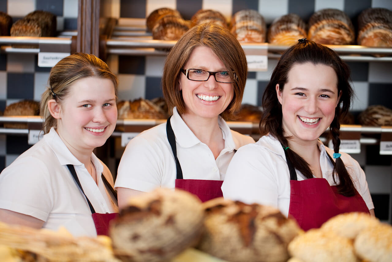 Betriebshaftpflicht für Bäckerei Konditorei Drei Bäckereifachverkäuferinnen in weißen Blusen und roten Schürzen lächeln vor Regalen voller Brote