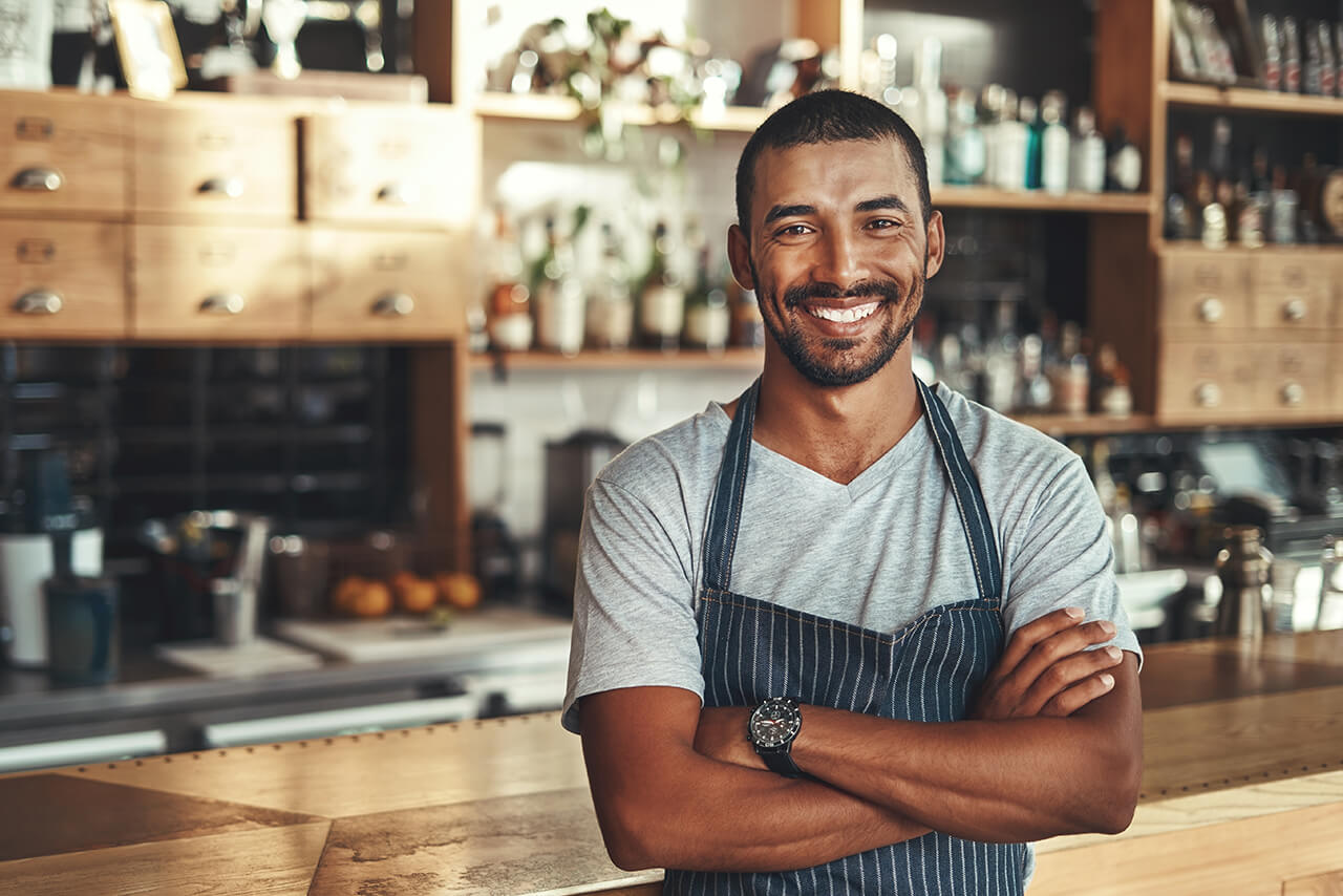 Betriebshaftpflichtversicherung für Café, Eiscafé und Eisdiele Lächelnder Barkeeper in grau gestreifter Schürze steht mit verschränkten Armen hinter der Theke einer Bar.