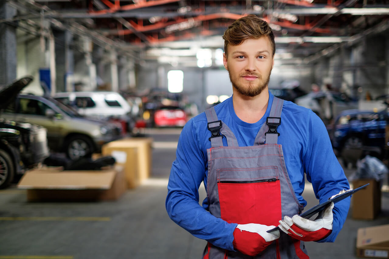 Kfz-Mechaniker in blauer Arbeitskleidung und Handschuhen hält ein Klemmbrett in einer Autowerkstatt.