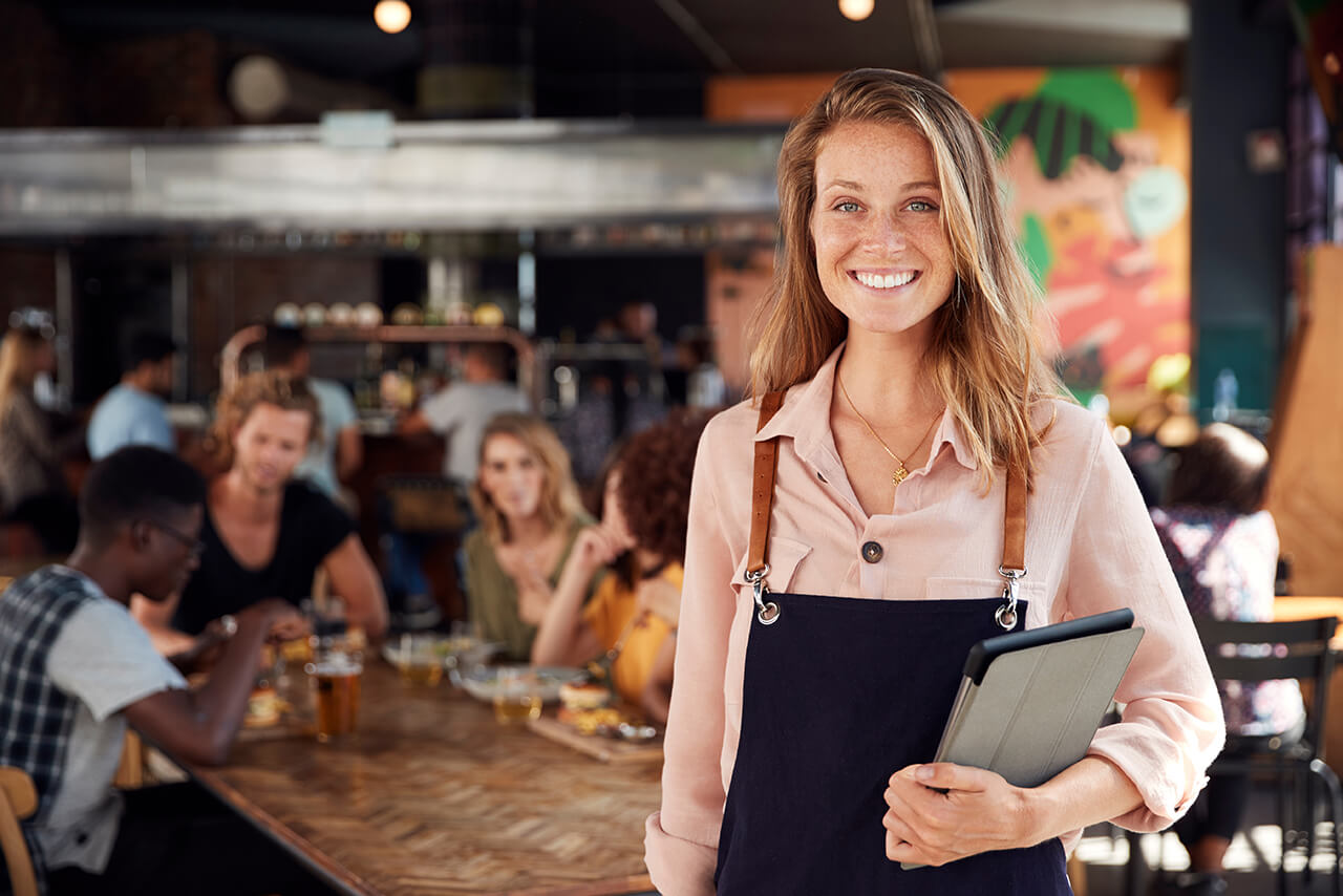Betriebshaftpflicht Restaurant | Online-Rechner Die Inhaberin eines Restaurants steht freudestrahlend in ihrem gut besuchten Restaurant und schaut uns an.