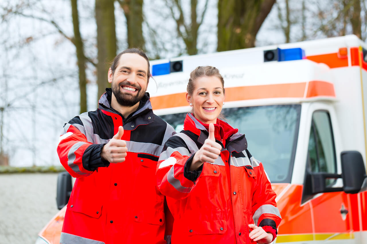 2 Rettungssanitäter im Einsatz - stehen vor Ihrem Einsatzfahrzeug und geben Daumen hoch Zeichen